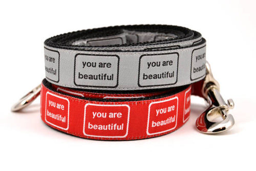 Two dog leashes stacked.  One is red with the words you are beautiful in white.  One is silver with the words you are beautiful in black.